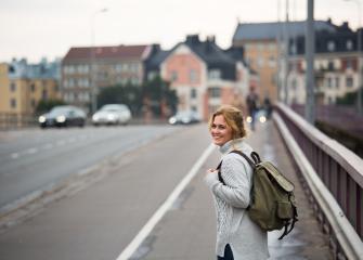 Valokuvassa nainen, jolla on reppu selässä, kävelee siltaa kaupungissa ja katsoo hymyillen taakseen.