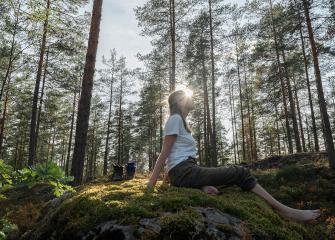 En person sitter barfota på en mossklädd klippa i en solig tallskog. Solljuset silas mellan träden.