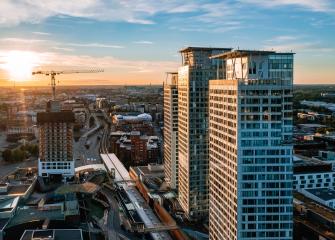 Ilmakuva kaupungista. Etualalla näkyy kolme korkeaa rakennusta ja yksi, jota vasta rakennetaan. Taustalla on liikennettä, nostokurki, kaupunkimaisemaa ja auringonlasku.