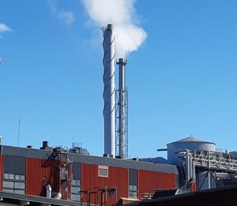 Kaksi tehtaanpiippua, josta tulee valkoista savua, taustalla on kirkas sininen pilvetön taivas.