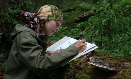 Metsässä ison lahopuun ääressä tutkija kirjoittaa tekemänsä lajihavainnot paperille.