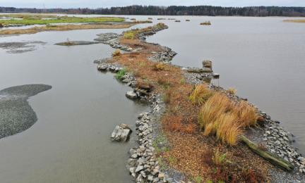 Kuvassa pitkänmallinen kiviaineksesta rakennettu saari, jolla kellastunutta syksyistä ruohokasvillisuutta. Ympärillä vettä ja taka-alalla toinen rakennettu saari.